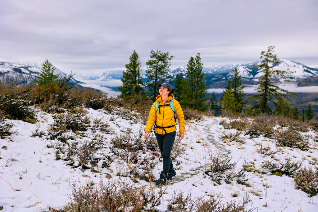 A woman hiking in winter with a jacket, backpack, and microspikes on her feet. Snow and mountains are in the background. 