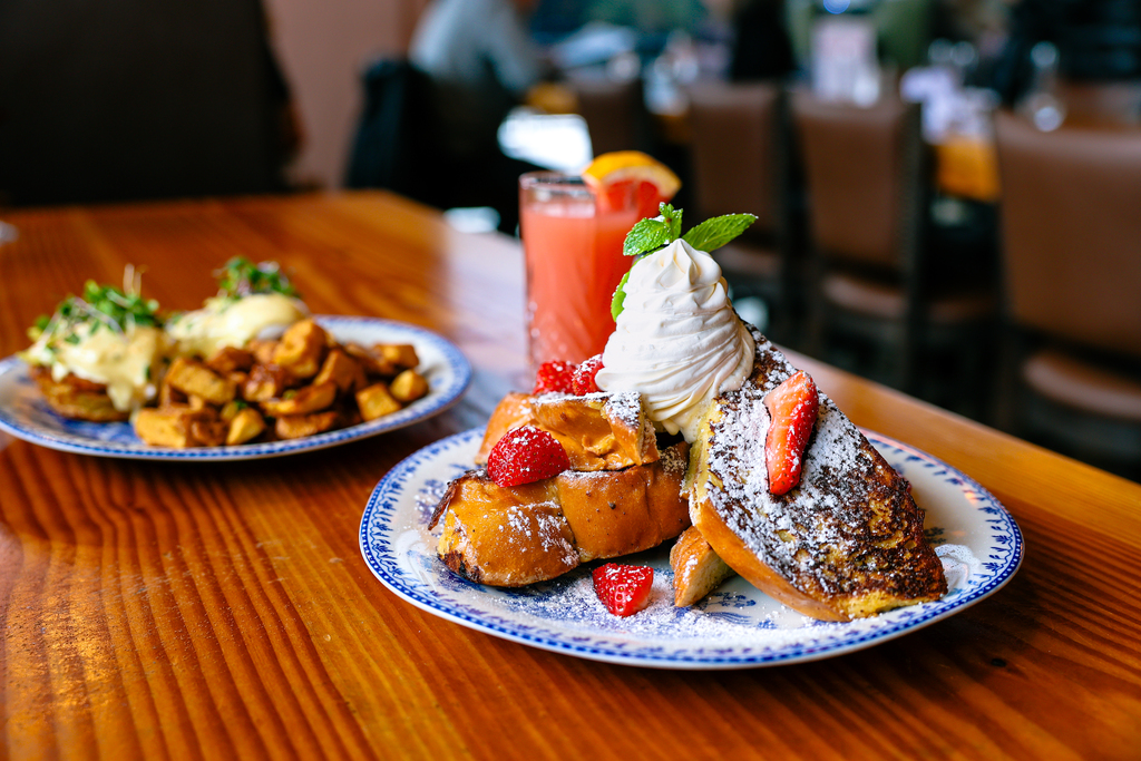 This picture shows two brunch entrees at Cecilia. The entrée in the foreground shows a giant stack of French toast with whipped cream on top. The Second entrée in the background shows eggs benedict with potatoes. There is also a glass of oink grapefruit juice in the background. Located near Vancouver, Washington.