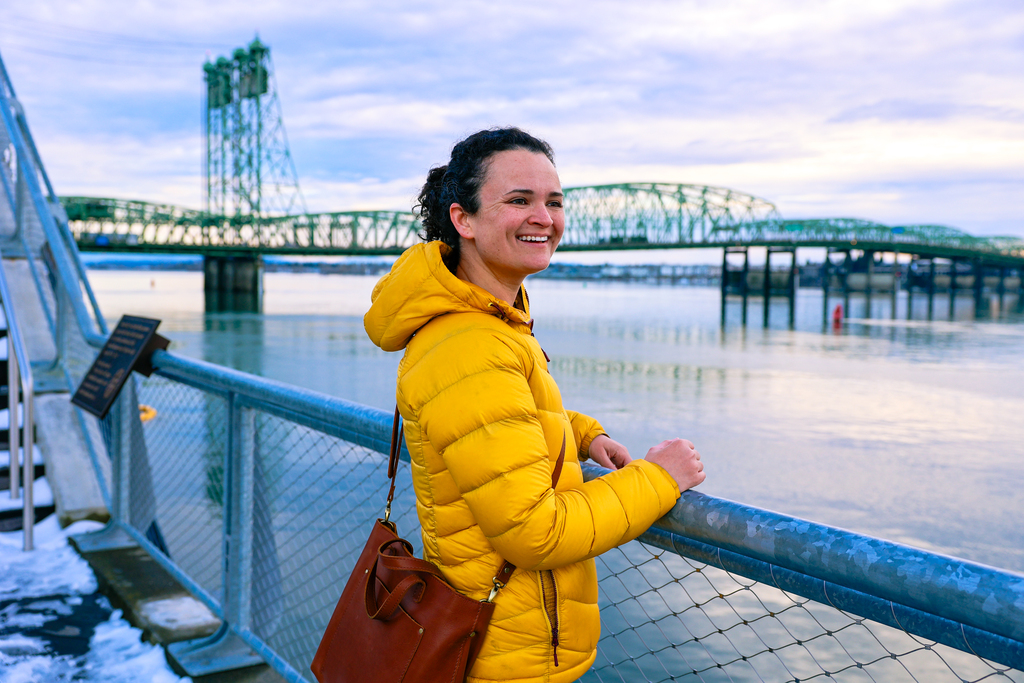 A woman in a yellow jacket standing on the Vancouver Waterfront overlooking the Columbia River. There is a green bridge in the background. In true Pacific Northwest fashion, the sky is cloudy. This is located near Vancouver, Washington.