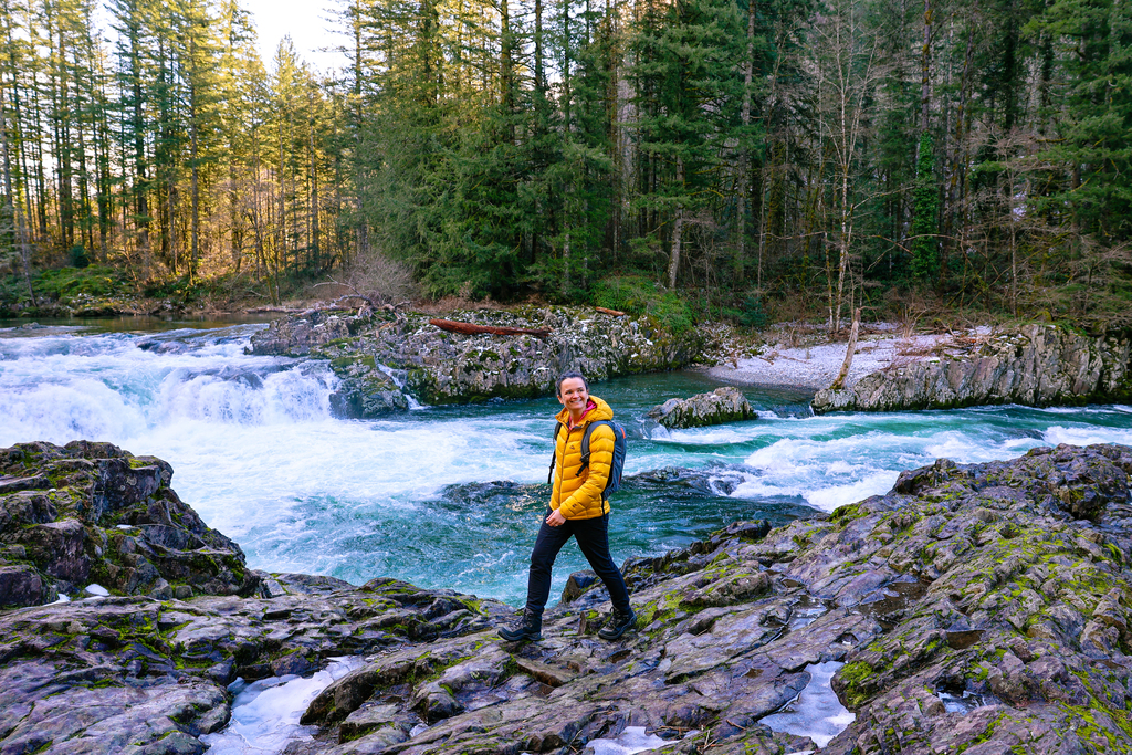 A woman in a bright yellow jacket hiking along a river bank next to a waterfall along the East Fork Lewis River in Vancouver, Washington.