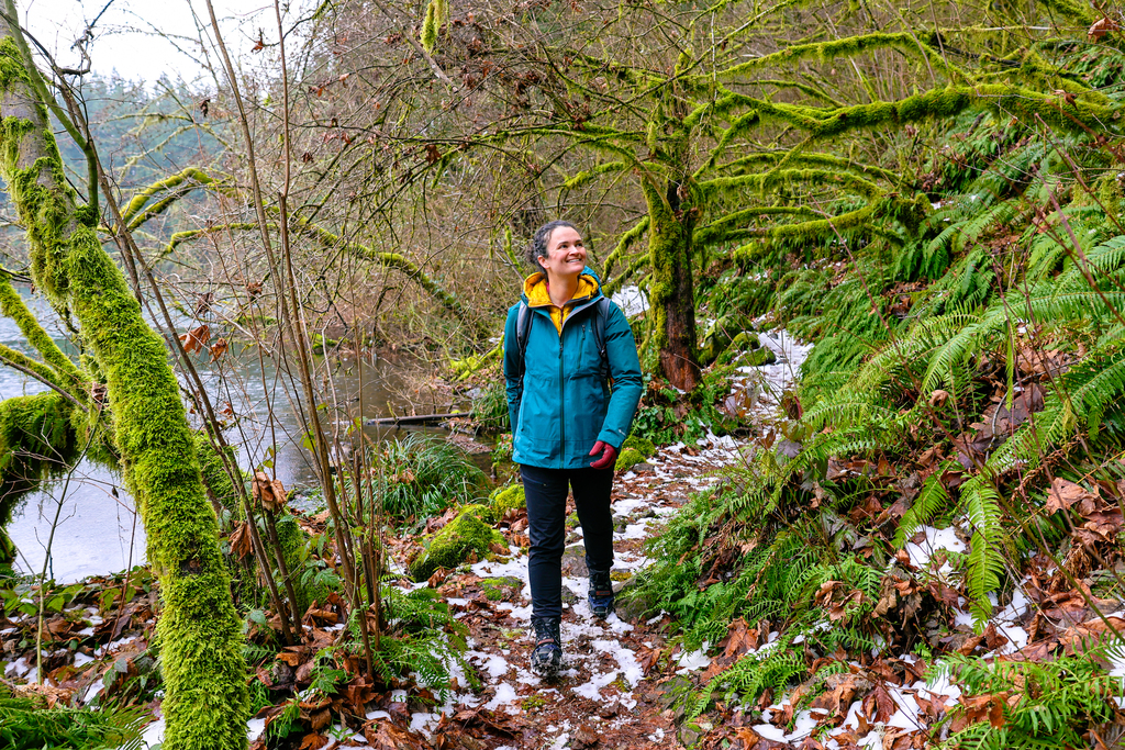 A woman in a teal rain jacket hiking along a hiking trail next to a lake. Green moss covers the surrounding trees. This hike is located near Vancouver, Washington.