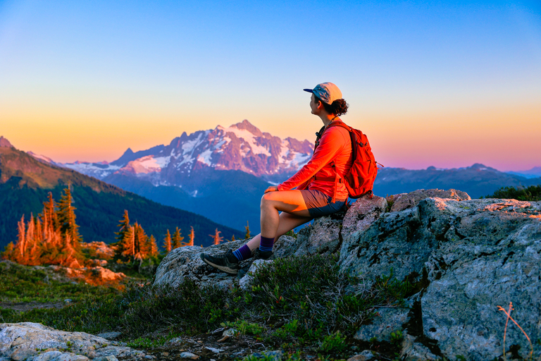 Woman sitting looking at Mount Shuksan and Mount Baker at sunset while on a summer hike at Yellow Aster Butte Trail in Washington state