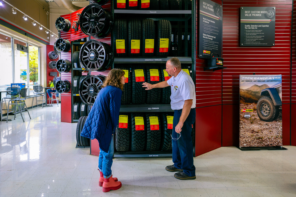 A lady and a man standing in front of a rack of tires. They are standing inside in the Les Schwab tire center. The man, a tire technician, is showing the lady a variety of tires on the rack, including the Les Schwab Open Range A/T Plus tires.