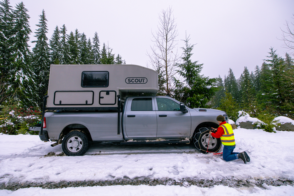 A picture of a parked grey RAM 3500 truck with a Scout truck camper in the snow. A lady wearing a yellow safety vest is installing tire chains on the front tires.